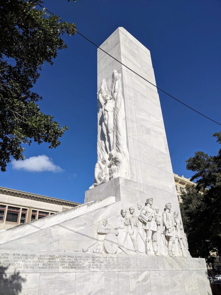 The tall white marble Alamo Cenotaph on Alamo Plaza, carved with figures of the defenders, against a deep blue sky