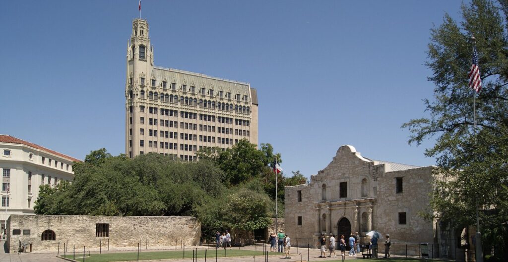 The Alamo church facade beside the long stone wall of the grounds, with the tall Emily Morgan hotel tower rising behind