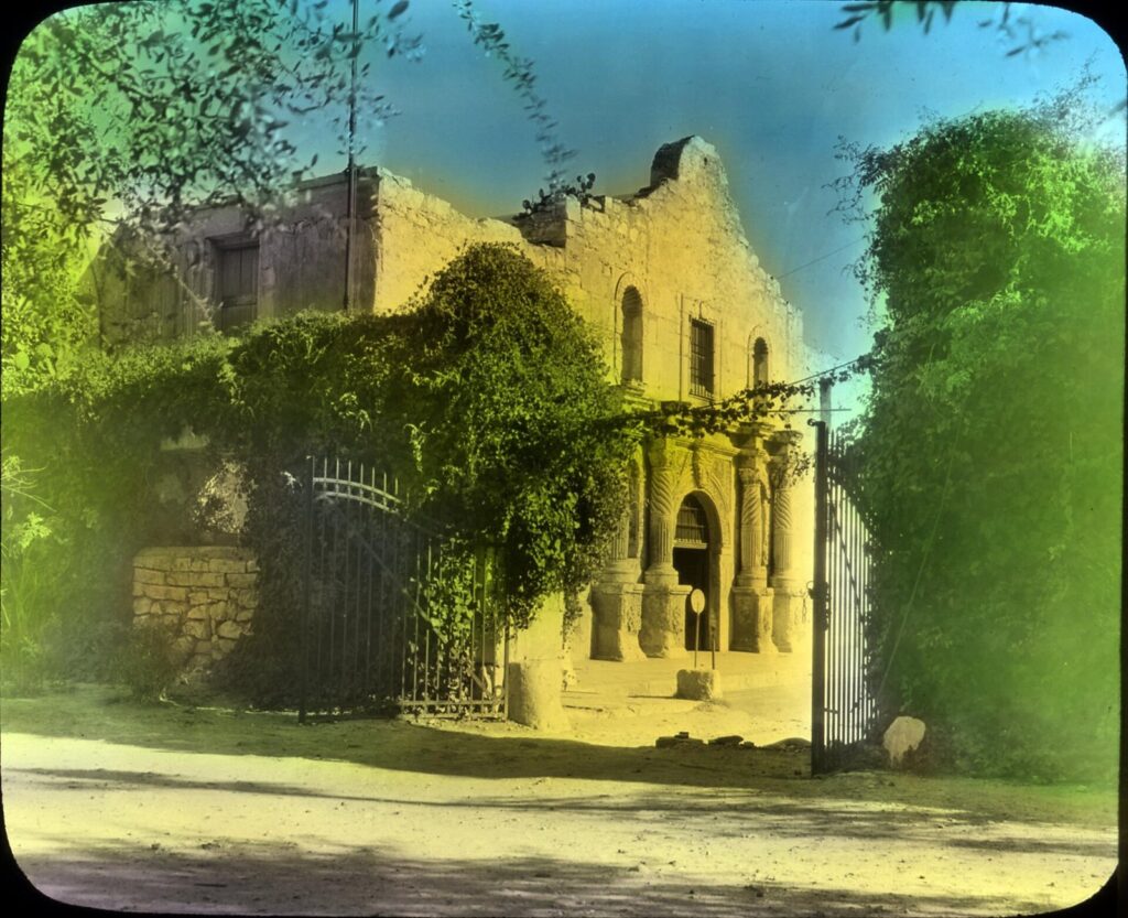 A hand-colored lantern slide of the Alamo church facade, partly covered in vines, with an iron gate at left