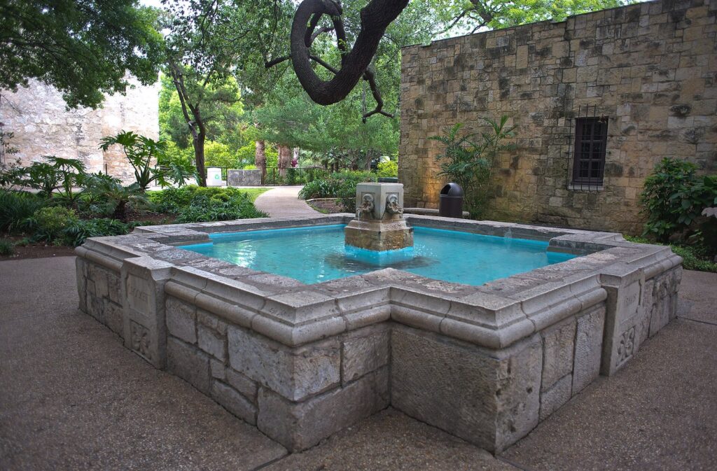 A square stone fountain with turquoise water in the Alamo Gardens, beside a weathered limestone wall and shade trees