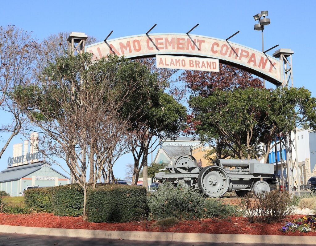An arched Alamo Cement Company sign over an old tractor and shrubs at the Alamo Quarry Market