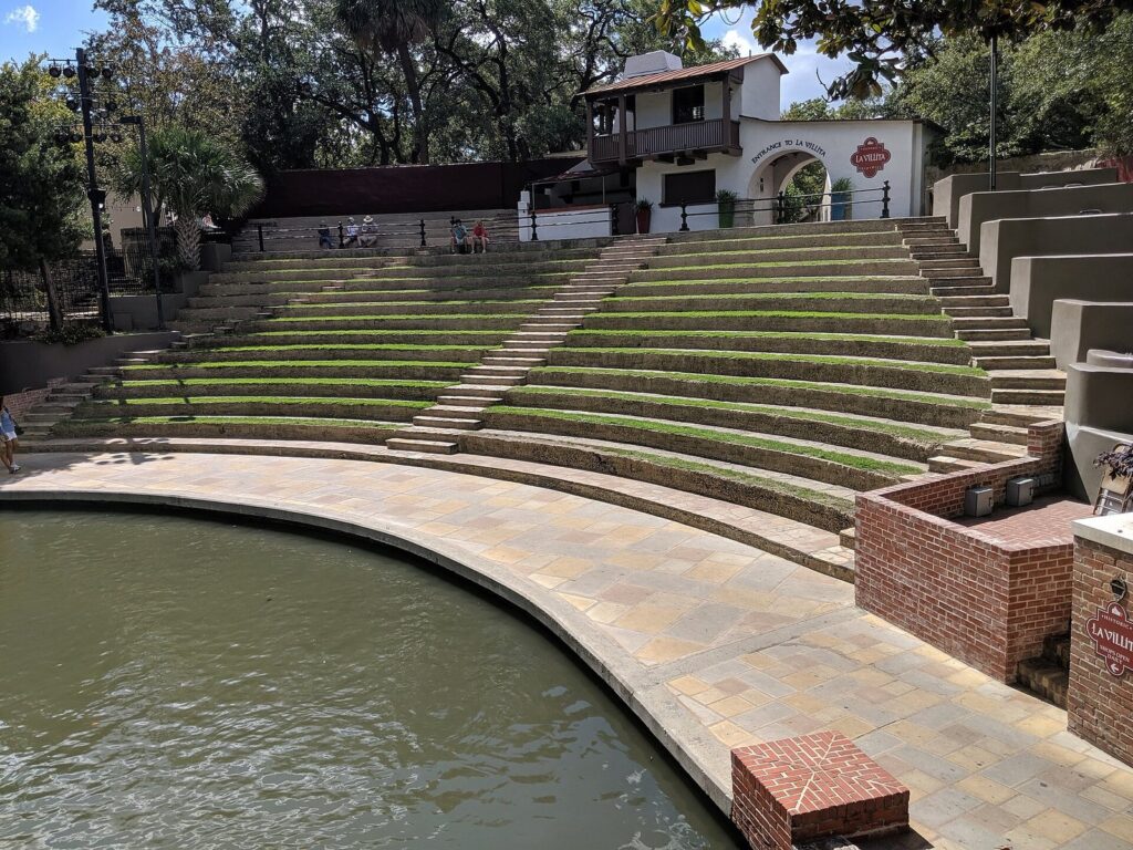 Grass-covered stone steps of the Arneson River Theatre seating curving down to the river below a white La Villita building