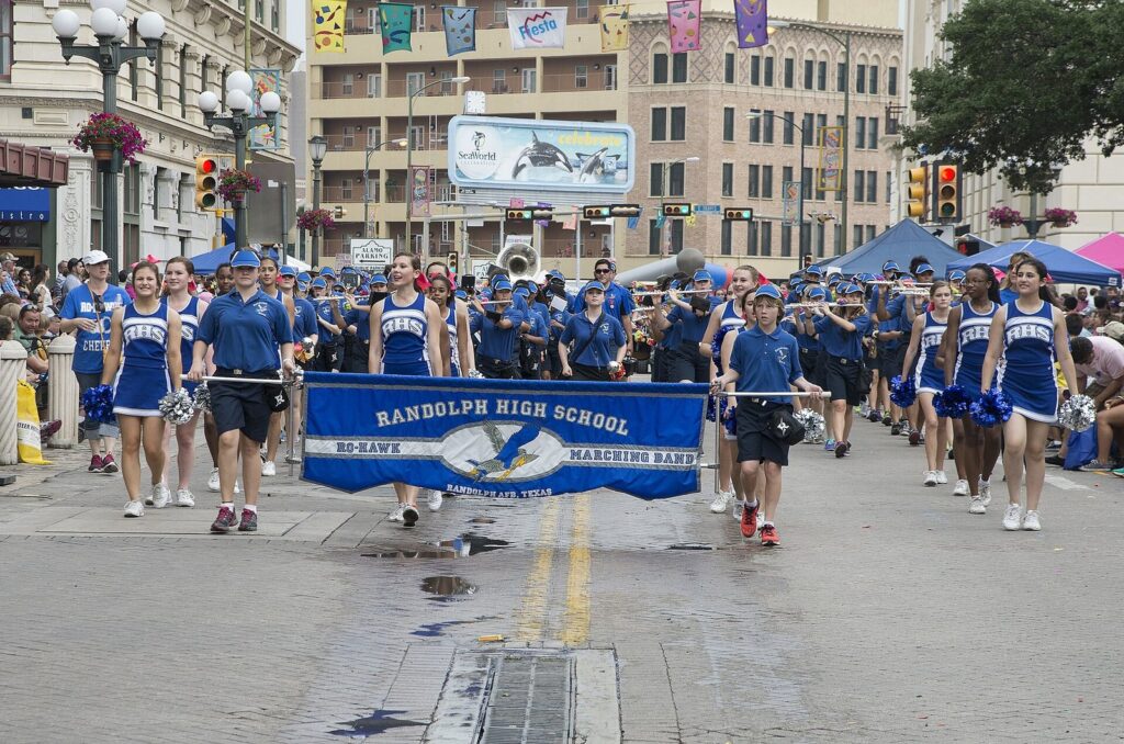 A high school marching band in blue carrying a banner down a downtown San Antonio street during the Battle of Flowers Parade