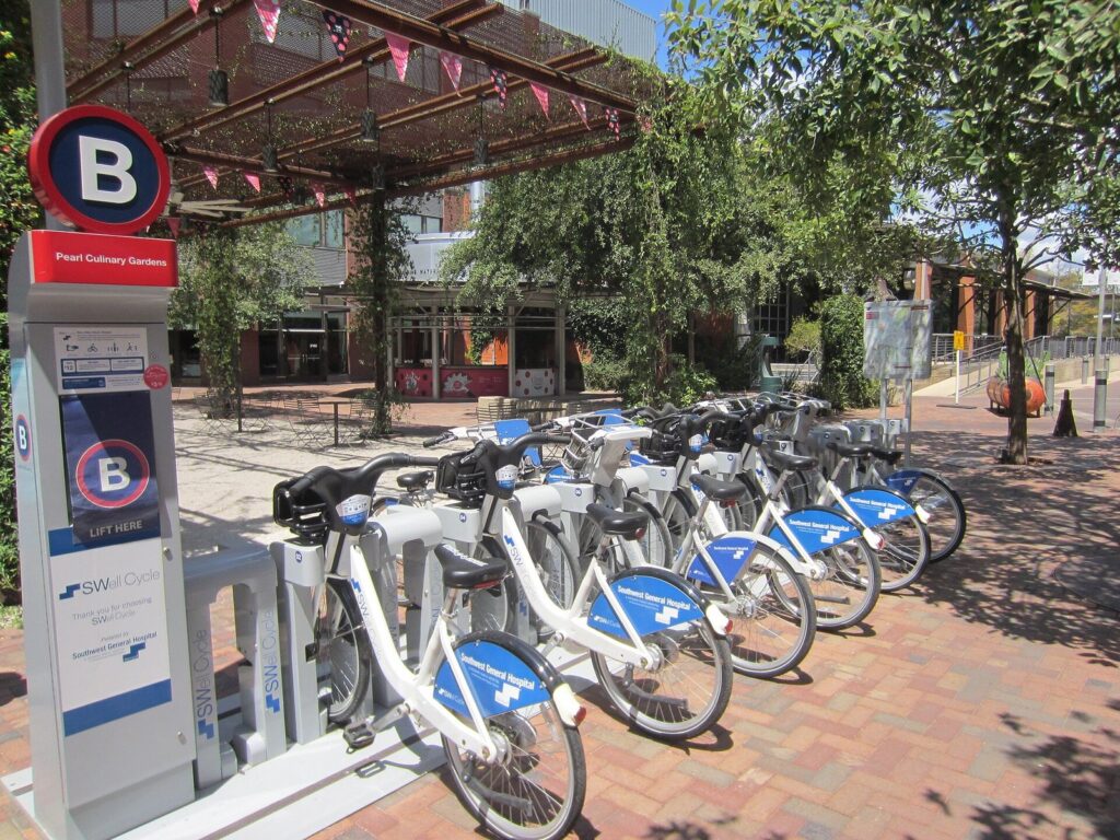 A row of BCycle bike share bikes docked at a kiosk under shade trees at the Pearl
