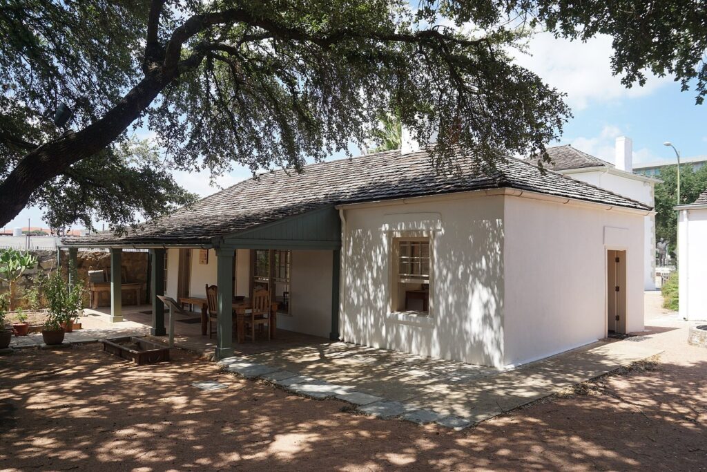 The low whitewashed adobe and limestone Casa Navarro under a spreading live oak, with a shaded porch and garden beds