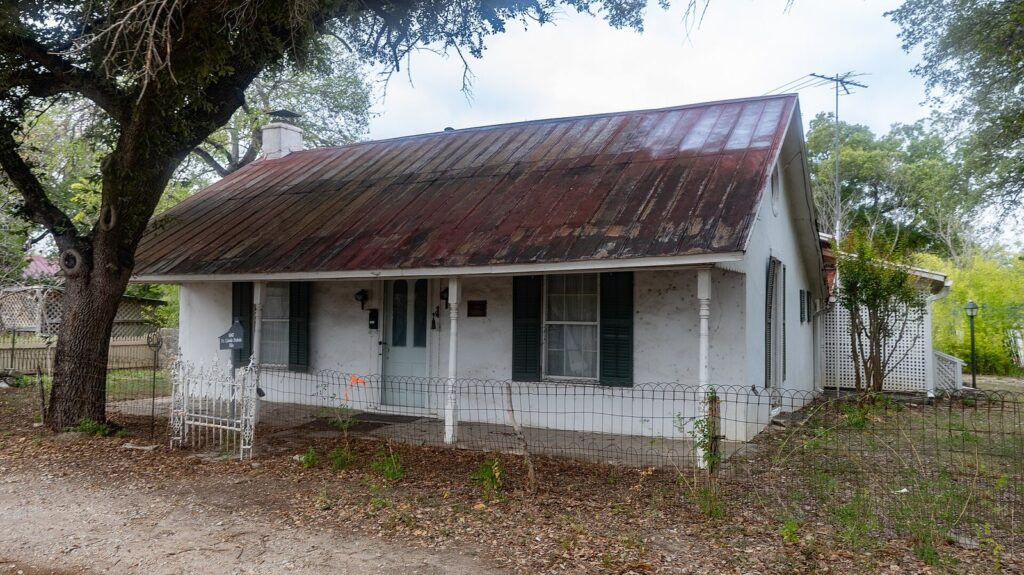 A small white Alsatian-style house with a steep rusted metal roof and green shutters under an oak tree in Castroville