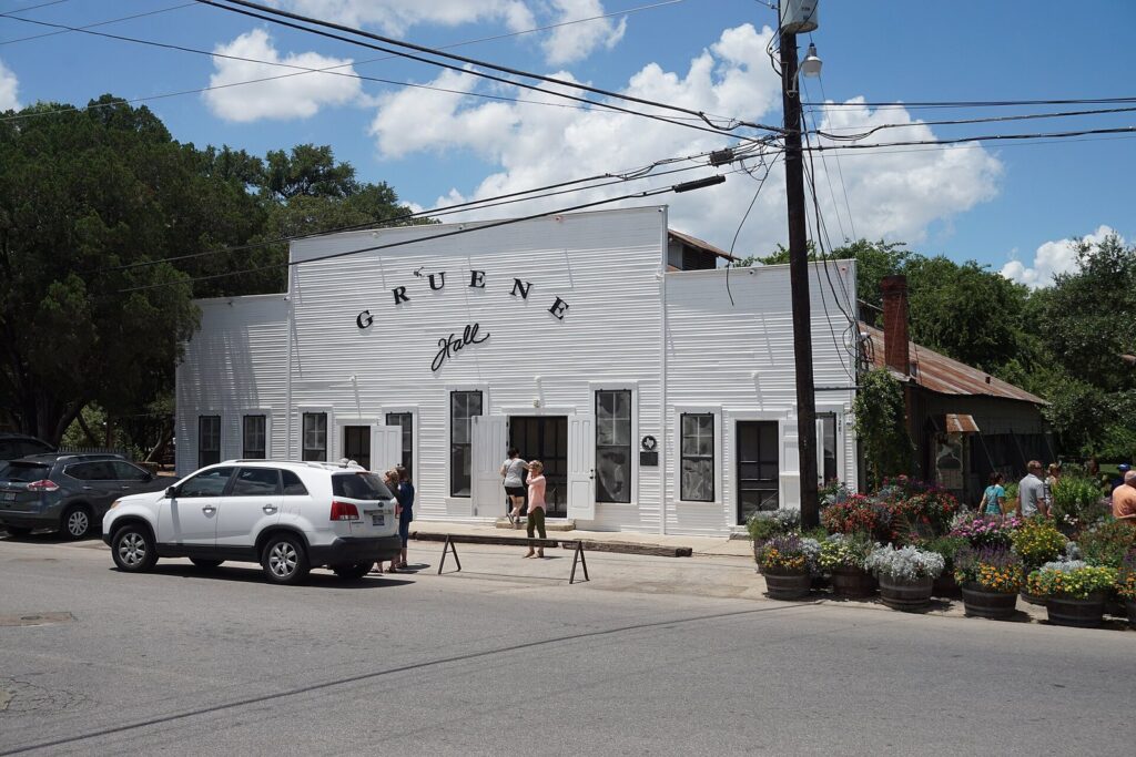 The white clapboard front of Gruene Hall with its arched painted sign, flower barrels and parked cars under a blue sky