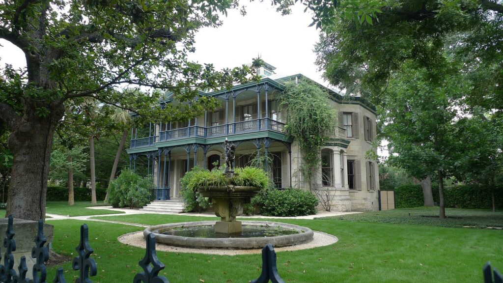 A two-story 19th-century house with iron-railed galleries and a round fountain on a lawn in the King William Historic District