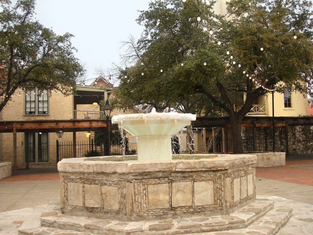 A round stone fountain in a paved courtyard at La Villita, with old limestone buildings and string lights in the oak trees behind