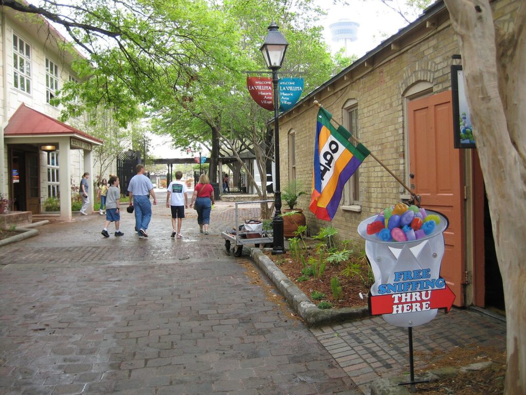 Visitors strolling a brick lane between old stone buildings and shade trees in La Villita