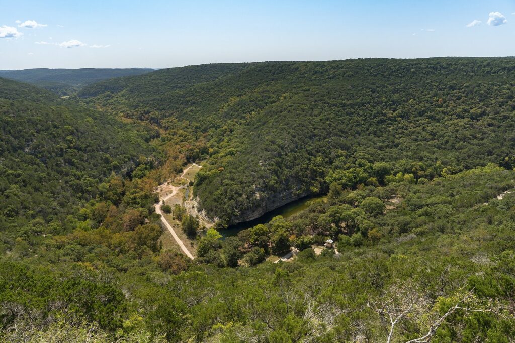 View from an overlook at Lost Maples across green wooded limestone hills and a canyon with a trail and pond below