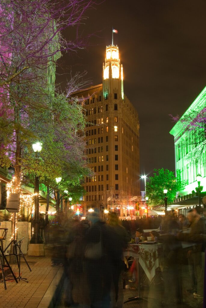 Crowds blurred in motion on a lit-up Houston Street at night during Luminaria, with the Emily Morgan Hotel tower behind