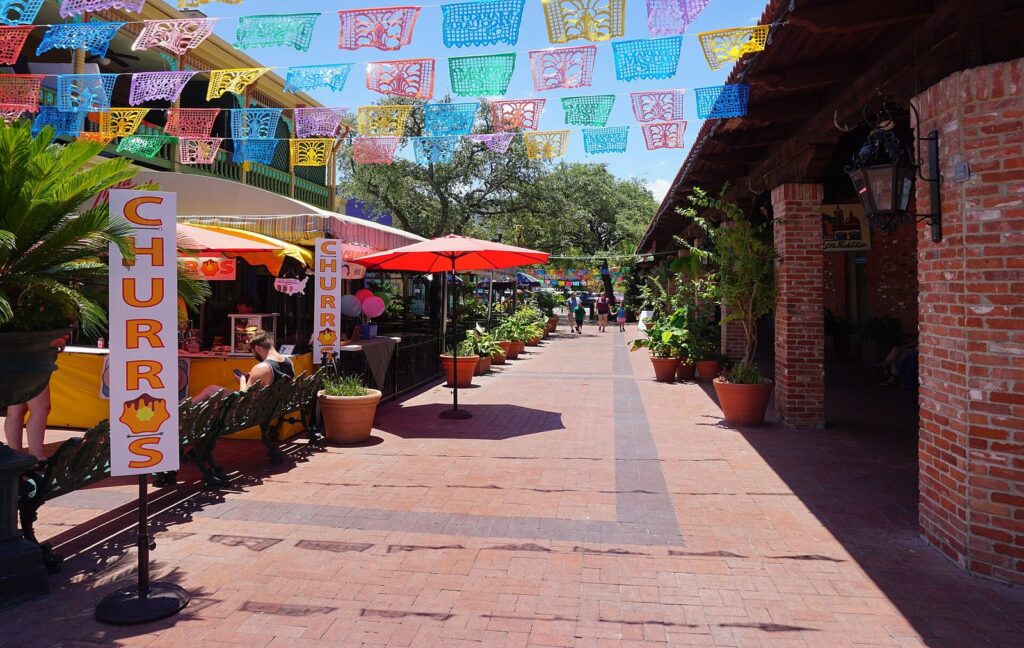 A brick walkway at Historic Market Square under rows of colorful papel picado, with a churro stand and potted plants, the heart of San Antonio shopping