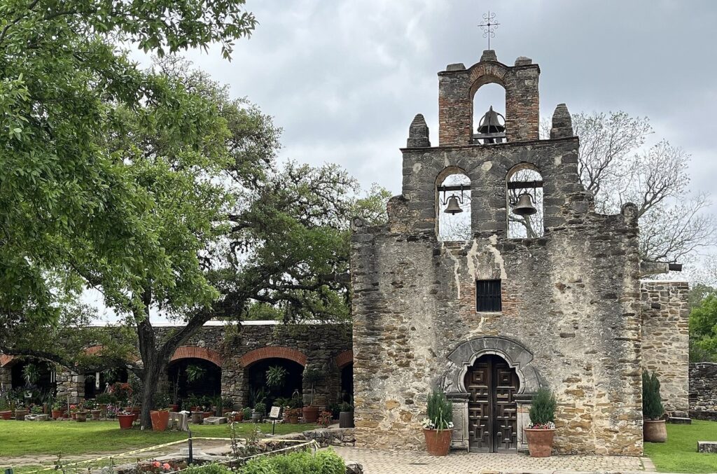The rough stone facade and bell wall of Mission Espada's church, with an arched doorway, beside green oak trees