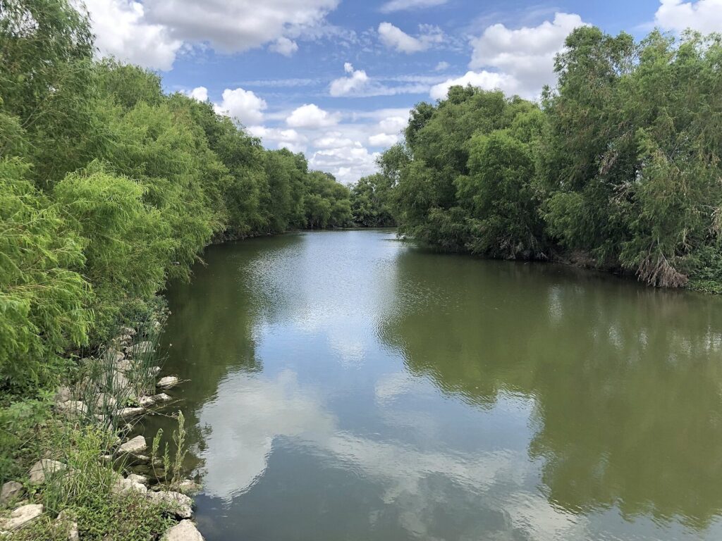 The San Antonio River on the Mission Reach between green brushy banks under a partly cloudy sky