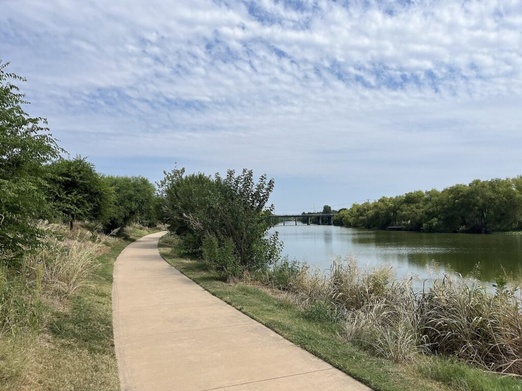 Paved hike-and-bike trail curving beside the San Antonio River on the Mission Reach under a cloudy sky