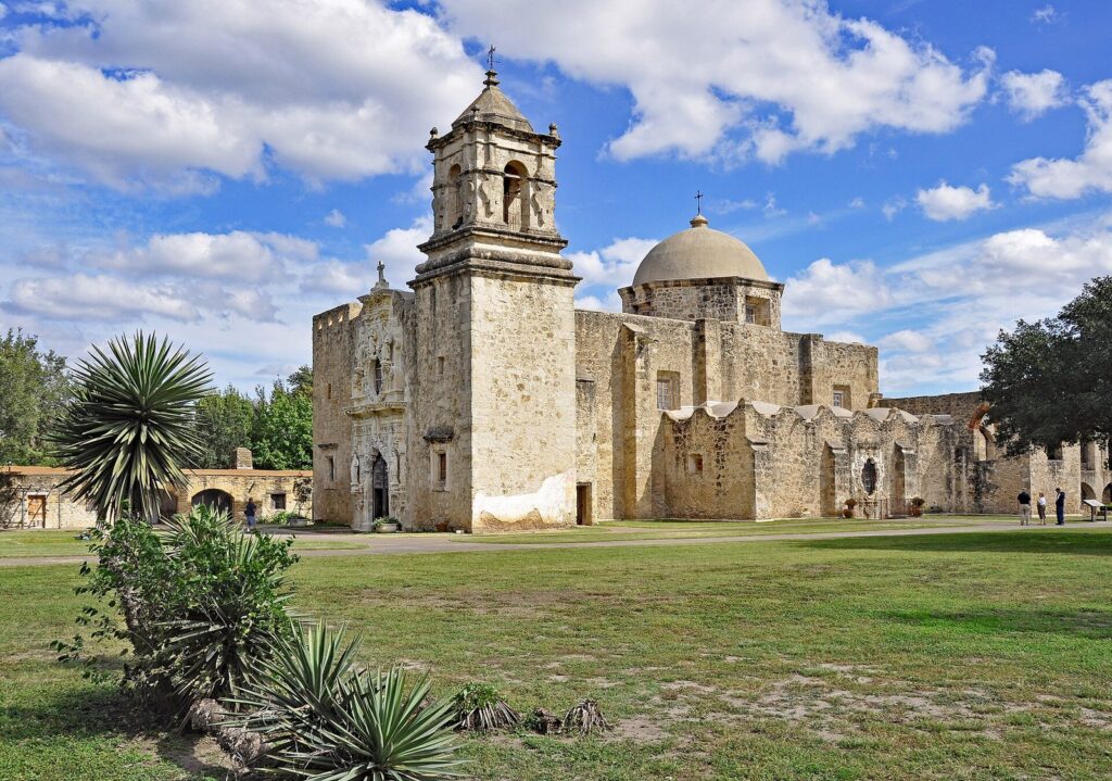 The limestone church, bell tower and dome of Mission San José under a blue sky with white clouds