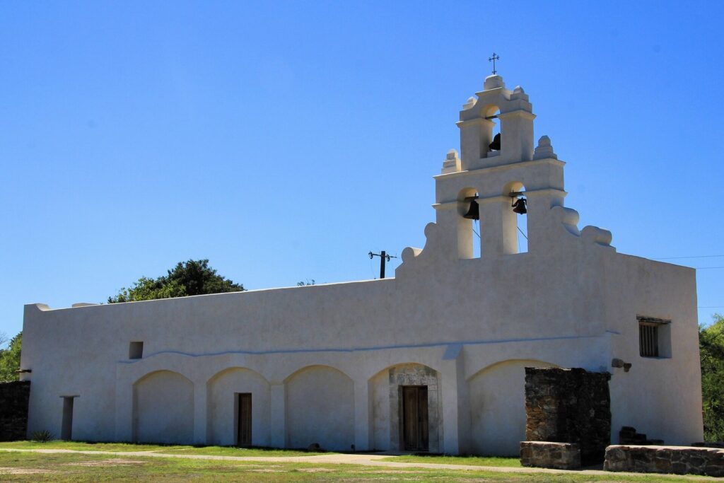 The white stucco church of Mission San Juan with its three-bell wall against a clear blue sky