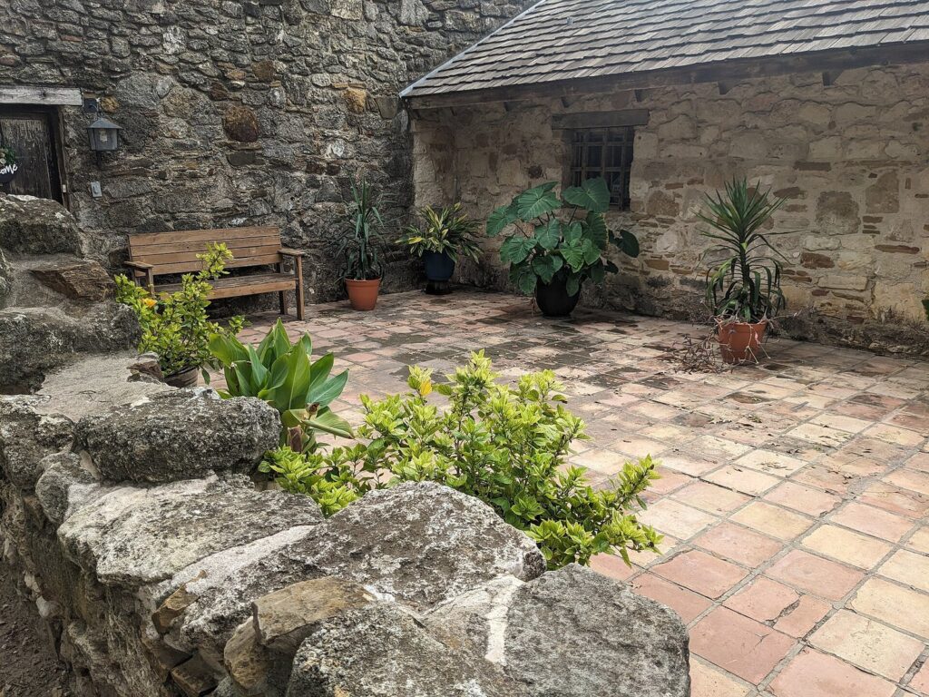 A shaded stone courtyard with potted plants, a wooden bench and old limestone walls at Mission San Juan Capistrano