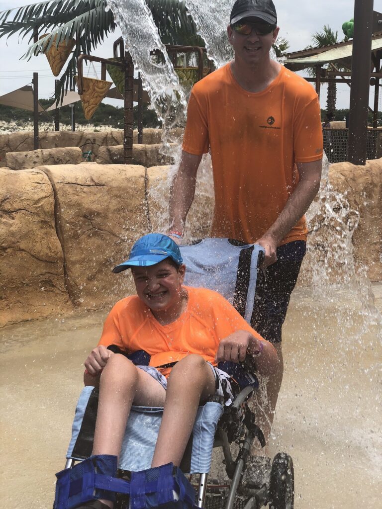 A smiling boy in a waterproof wheelchair pushed through a splash of water by a staff member at Morgan's Inspiration Island