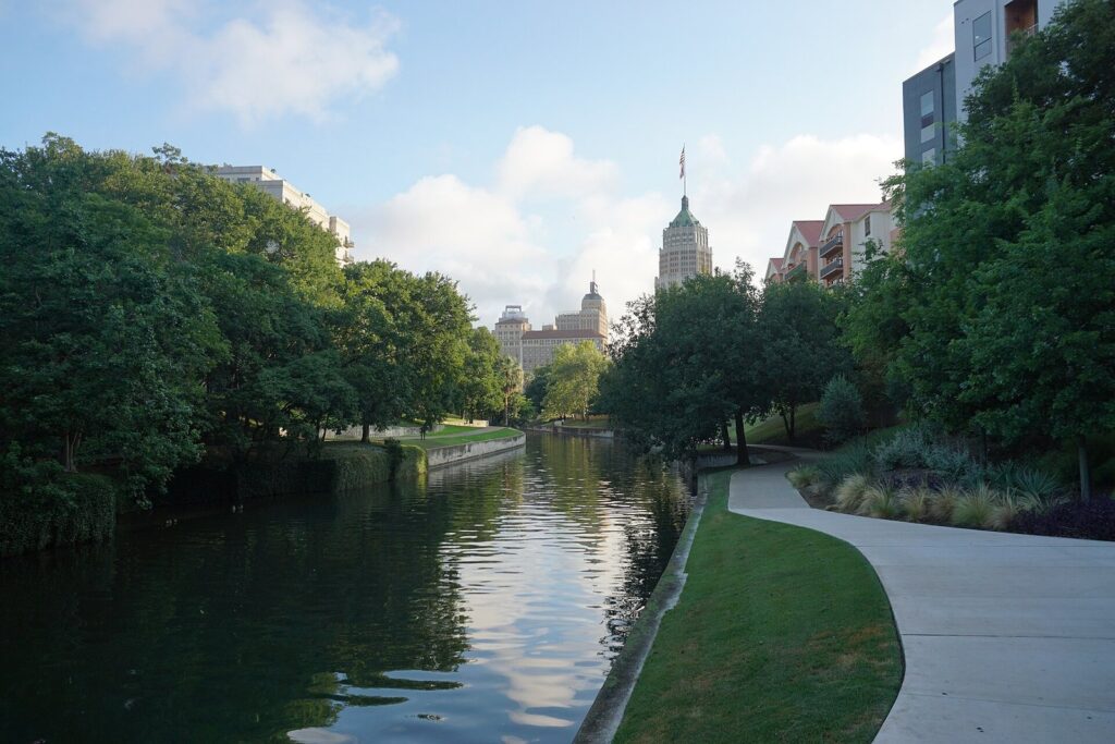 A curving paved path beside the calm San Antonio River on the Museum Reach, with green trees and downtown towers ahead