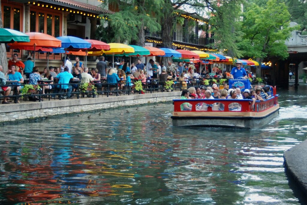 River Walk restaurant patios under rows of colorful umbrellas with a crowded tour boat passing on the water