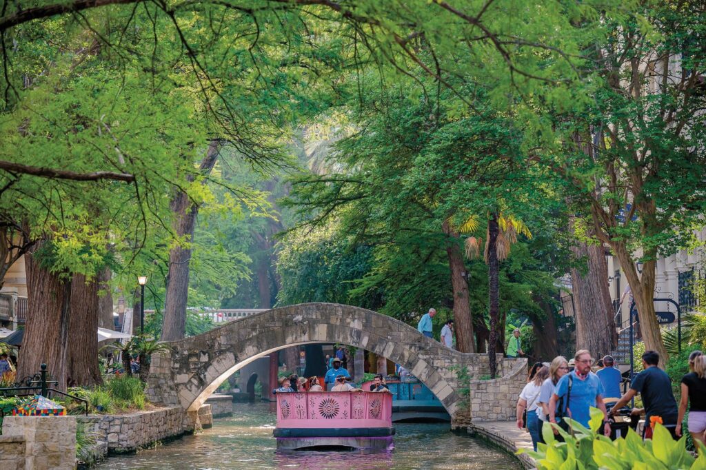 A tour boat passing under an arched stone footbridge on the River Walk as visitors watch from the shaded path, a classic first sight among things to do in San Antonio for first time visitors