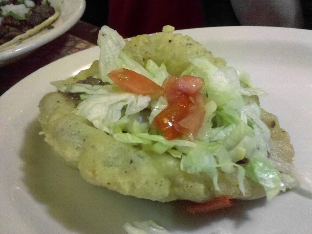 A puffy taco with a crisp, blistered masa shell topped with shredded lettuce and diced tomato on a white plate