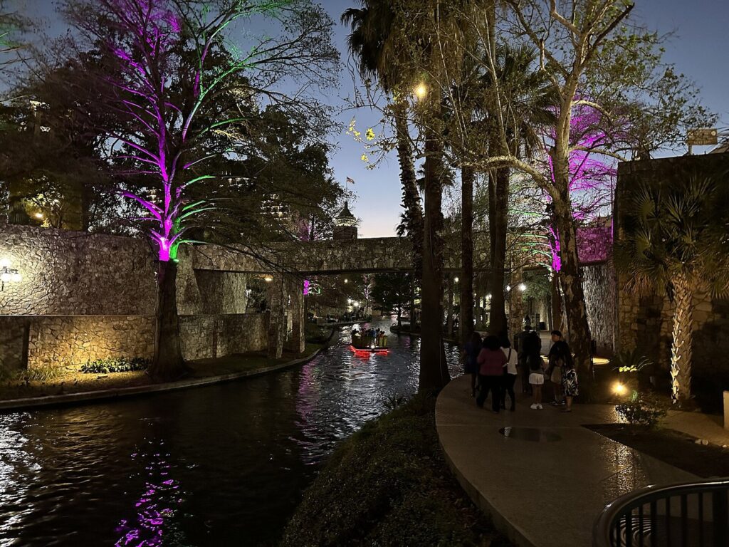 Walkers on the downtown River Walk at night beneath cypress trees lit purple, with a boat far down the river