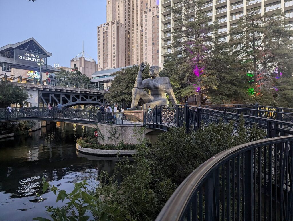 Footbridges over the San Antonio River Walk beside the Shops at Rivercenter, with a seated sculpture and hotel towers behind