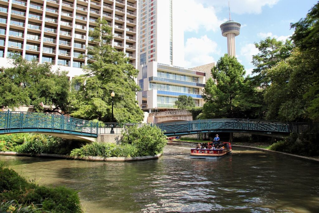 A tour boat passes under green footbridges on the San Antonio River Walk, with hotels and the Tower of the Americas behind