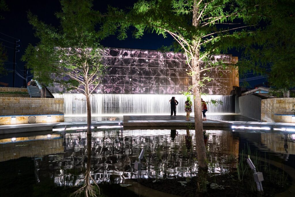 San Pedro Creek Culture Park at night, with a lit wall of water falling into a reflecting channel lined by trees