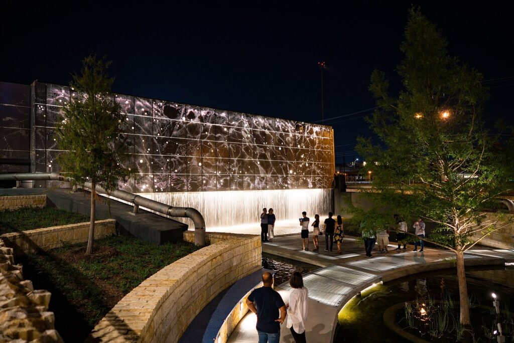Visitors walking along a lit paseo beside a long illuminated wall and waterfall at San Pedro Creek Culture Park at night