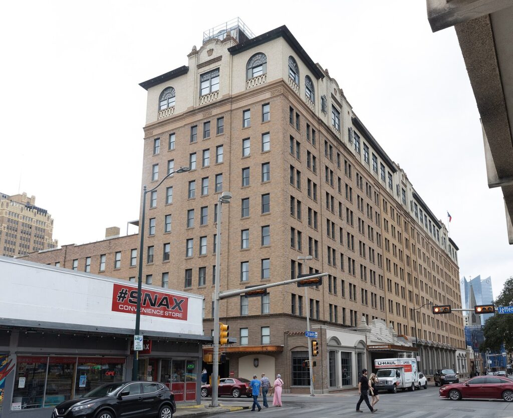 The tall tan brick St. Anthony hotel on a downtown San Antonio street corner, with a convenience store and pedestrians below