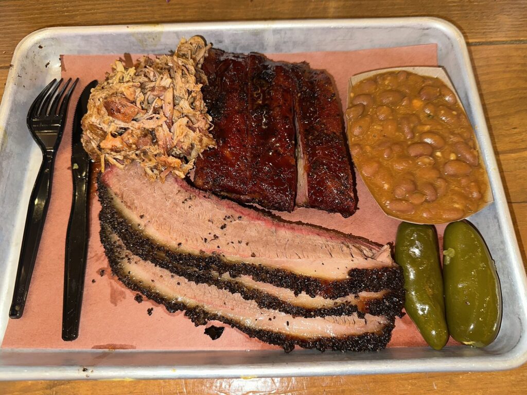 A metal tray of sliced smoked brisket with a dark bark, pork ribs, pulled pork, pinto beans and pickled jalapeños on butcher paper