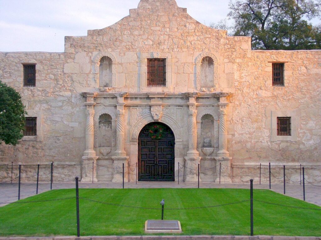 The limestone facade of the Alamo church in San Antonio in warm evening light, behind a green lawn and a low chain barrier