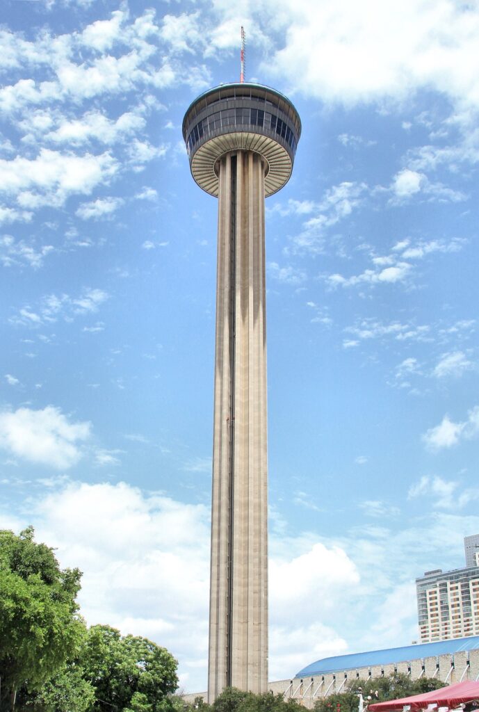 The Tower of the Americas rising above trees at Hemisfair against a blue sky with scattered clouds