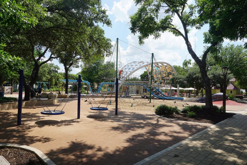 Swings, climbing nets and a play structure under shade trees at Yanaguana Garden playground in Hemisfair
