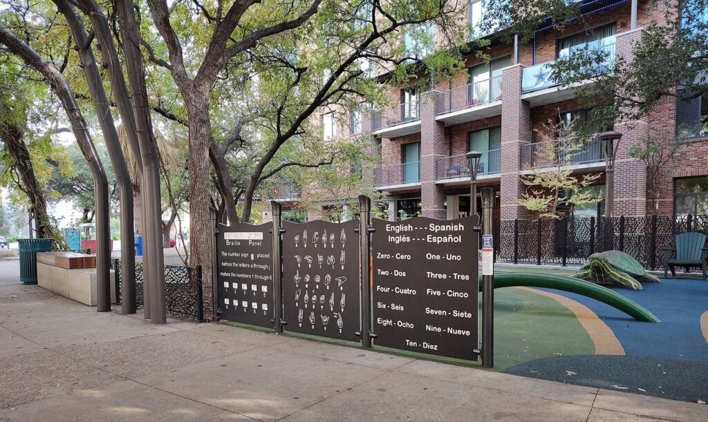 The Yanaguana Garden playground at Hemisfair with a bilingual counting panel, play surfaces and shade trees beside brick apartments