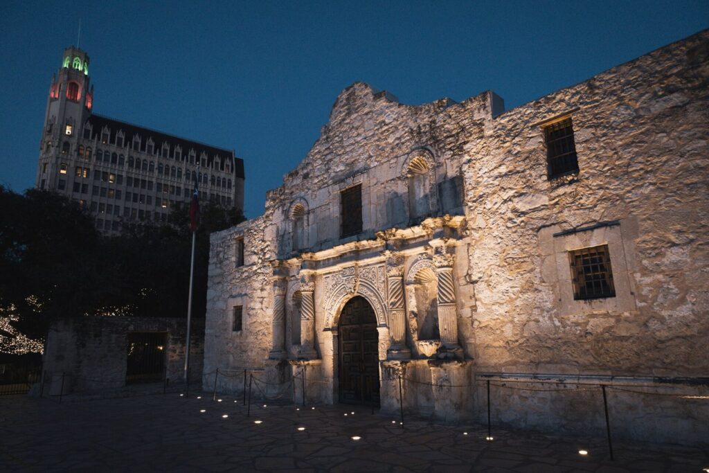 The Alamo church facade lit at dusk, with the Emily Morgan hotel tower rising behind it against a deep blue sky