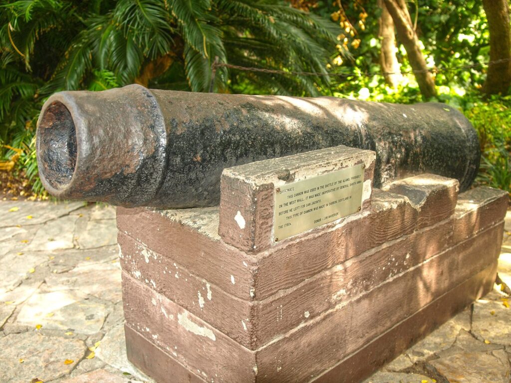 An old iron carronade on a wooden mount with a brass plaque, among palms and plants on the Alamo grounds