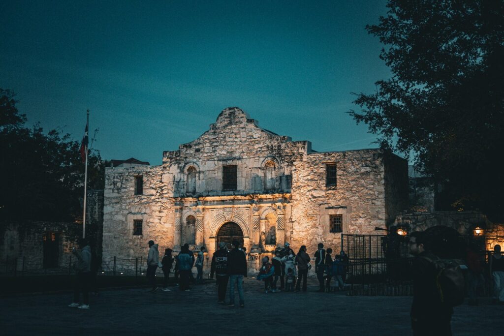 Visitors gathered on Alamo Plaza in front of the floodlit Alamo church at dusk, with trees framing the scene