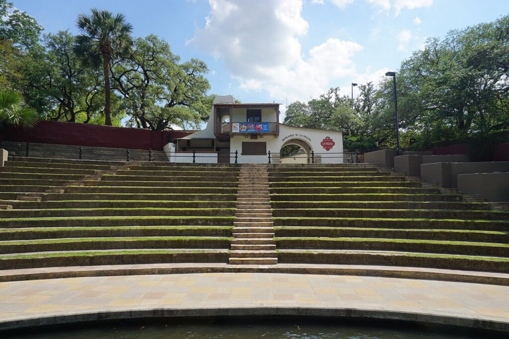 Grass-covered stone steps of the Arneson River Theatre curving down to the river, with a white La Villita building at the top