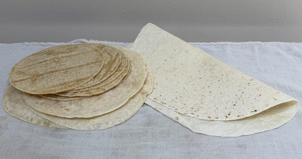 A stack of yellow corn tortillas on top of flour tortillas, with a large folded flour tortilla beside them on a white cloth