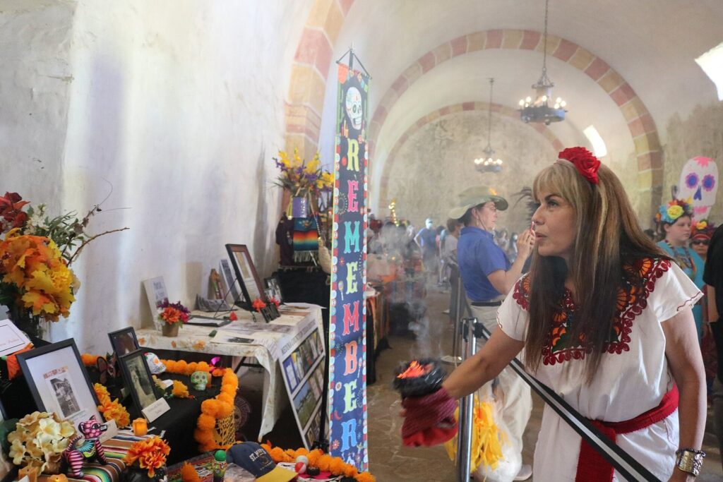 A woman in an embroidered white dress waves incense over a Day of the Dead altar with photographs and marigolds inside an arched hall