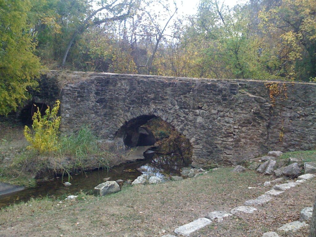 The stone Espada Aqueduct crossing a creek on low arches, with autumn trees behind