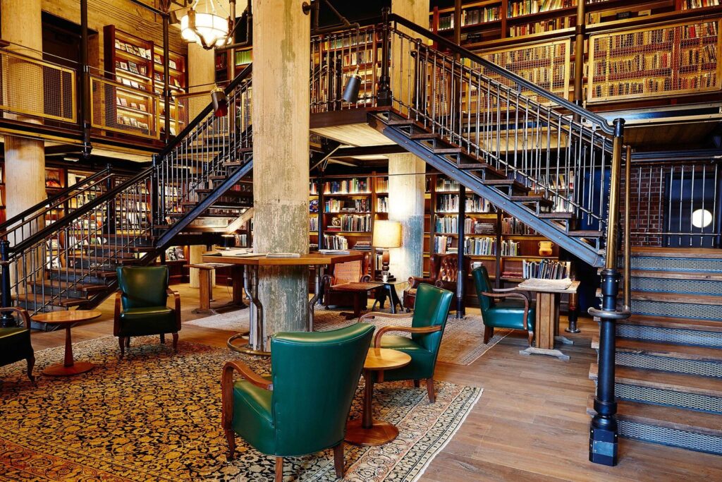 The two-story library at Hotel Emma, with iron staircases, tall bookshelves, green leather chairs and a patterned rug among old brewery columns