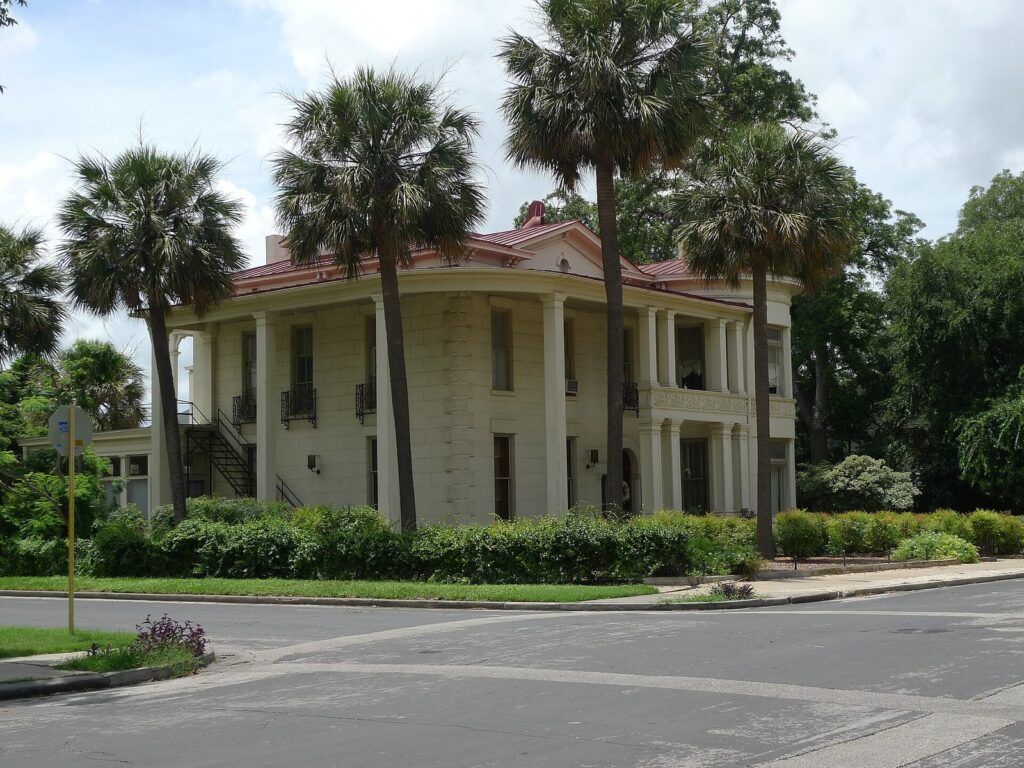 A two-story 19th-century stone house with deep porches and tall palm trees on a corner in the King William Historic District