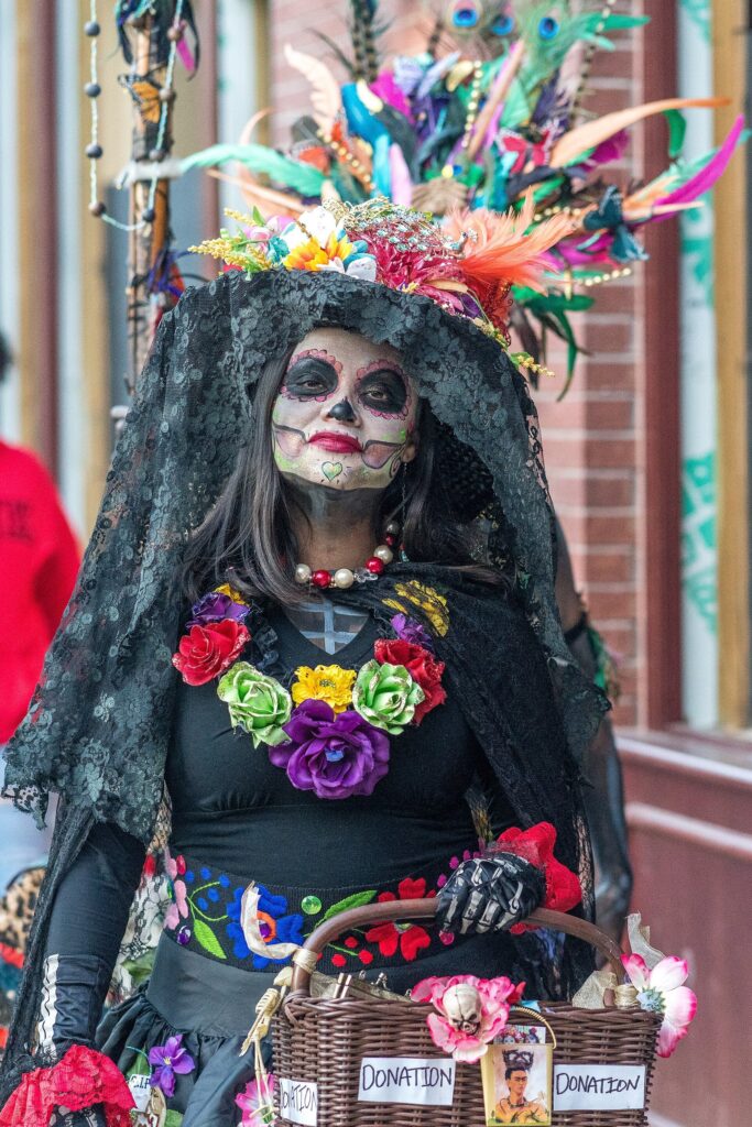 A woman dressed as La Catrina with calavera face paint, a black veil, a feathered floral headdress and a necklace of paper flowers