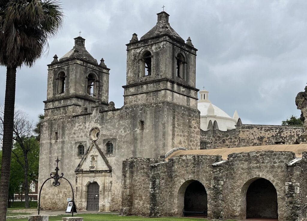 The twin bell towers, dome and weathered stone walls of Mission Concepción in San Antonio, beside a palm under a cloudy sky